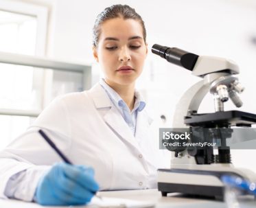 lab note2 Serious thoughtful young woman in lab coat sitting at table and making records about research in laboratory